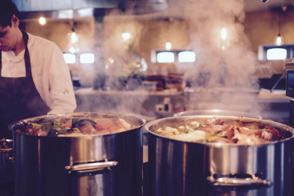 Chef working beside large steaming stock pots in a commercial kitchen